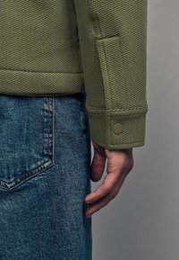 Kaki tekstureret jakke med synlige syninger og en trykknapmanchet, parret med denim jeans, der viser en baglomme.