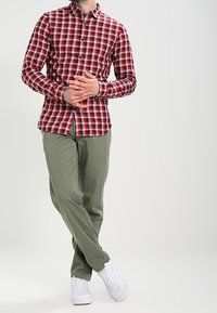 Mann steht mit verschränkten Beinen, trägt ein rotkariertes Hemd, olivgrüne Hose und weiße Sneaker, die Hände vor sich gefaltet.