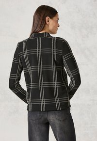Svart texturerad tröja med ett vitt rutmönster, långa ärmar och en bakvy med hög halsringning. Matchas med mörka jeans.