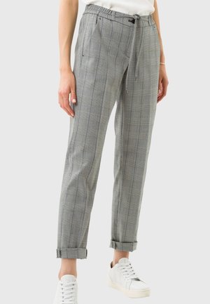 Femme portant un pantalon gris à carreaux retroussé, des baskets blanches et un haut blanc, debout devant un fond clair uni.