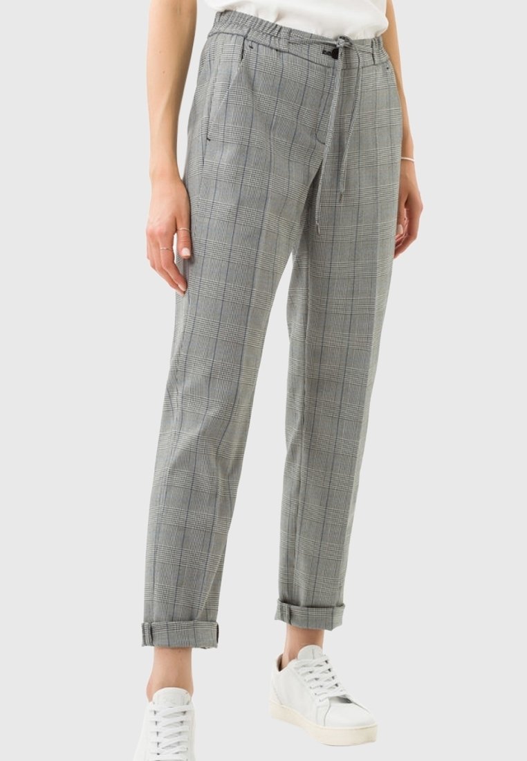 Femme portant un pantalon gris à carreaux retroussé, des baskets blanches et un haut blanc, debout devant un fond clair uni.