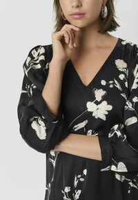 Blouse noire à fleurs avec un décolleté en V, des manches bouffantes et une texture satinée présentant des imprimés botaniques blancs et des poignets élastiques.