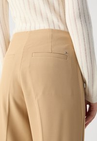 Beige teksturerede bukser med en skræddersyet pasform, der har baglommer og et lille logodetalje nær taljen. Delvist synlig cremet stribet sweater.