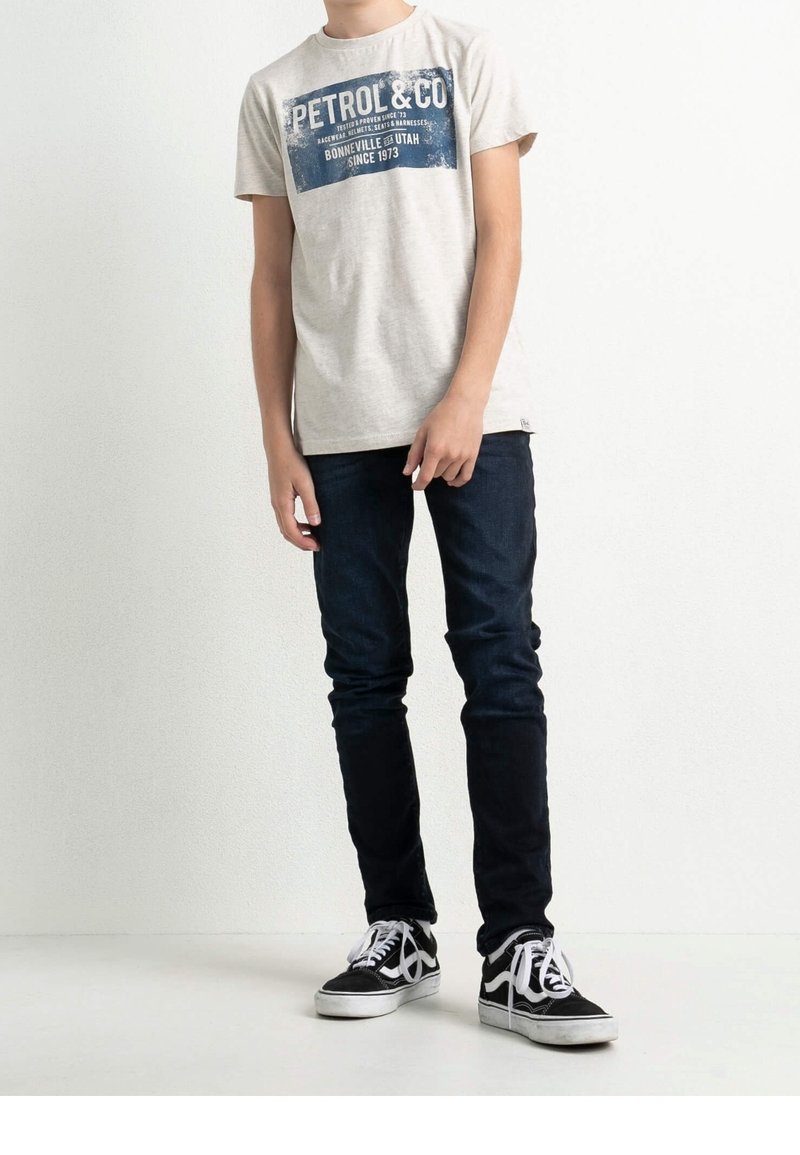 Camiseta gris claro con estampado gráfico azul, jeans ajustados oscuros, zapatillas negras y blancas con cordones. Diseño simple y apariencia casual.