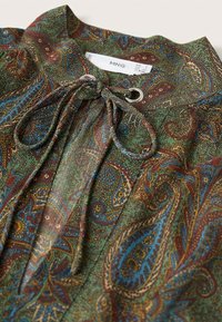 Blouse à motifs avec une base verte ornée de motifs paisley rouges, bleus et dorés. Comprend une fermeture par nouage et des éléments décoratifs en métal.