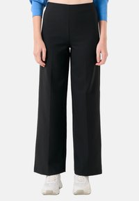 Pantalons larges noirs en tissu lisse et texturé, taille haute et lignes épurées, associés à des chaussures de sport blanches.