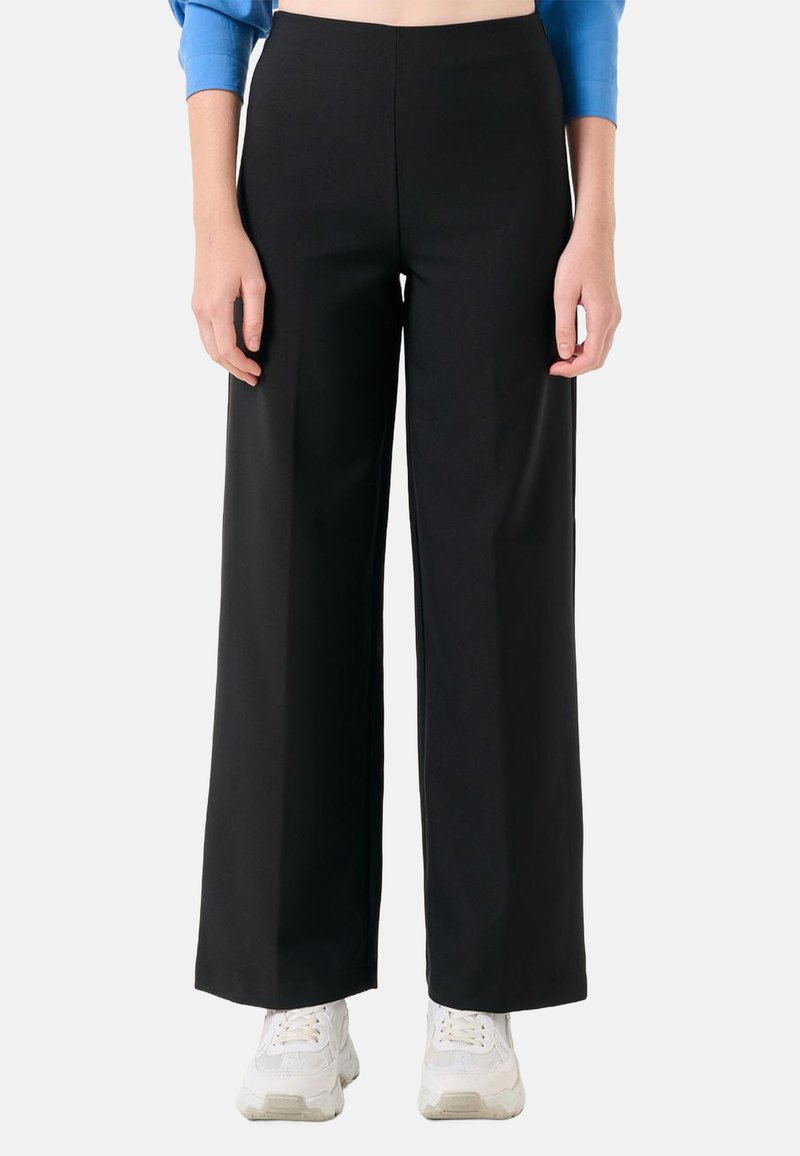 Zwarte wijde broek van een gladde, textiele stof, hoge taille en strakke lijnen, gecombineerd met witte sportschoenen.