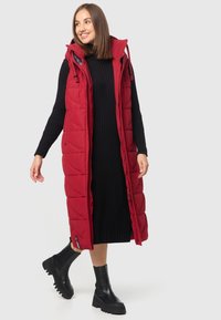 Gilet long rembourré rouge avec capuche, doté d'une fermeture éclair et de poches latérales, superposé sur une robe côtelée noire et des bottines noires.