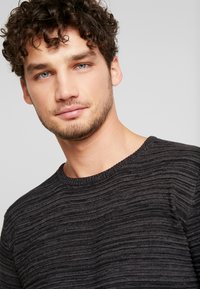 Dark gray knit sweater with a textured, striped pattern and a round neckline, worn by a male model with curly hair and facial stubble.