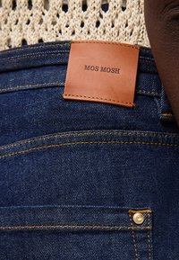 Κοντινή λήψη σκούρων μπλε τζιν με δερμάτινο ταμπελάκι ταμπά με την ένδειξη "MOS MOSH" και κίτρινες ραφές.