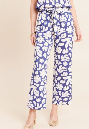 Frau trägt blaue Hosen mit weitem Bein, weißem abstraktem Blumenmuster und einem gebundenen Gürtel, kombiniert mit goldfarbenen High-Heels und einem goldenen Armband.