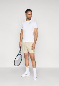 Ανδρική αθλητική ενδυμασία τένις αποτελούμενη από λευκό κοντομάνικο πουκάμισο, μπεζ σορτς, λευκά παπούτσια Nike και κρατώντας ένα ρακέτα με μπλε λεπτομέρειες.