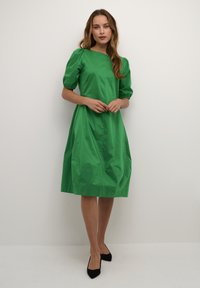 Rochie din bumbac verde, cu mâneci scurte bufante, croială dreaptă și tiv la nivelul genunchilor. Textură netedă, cu un design simplu și fără modele vizibile.