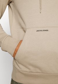 Sudadera beige con bolsillo canguro, puños acanalados y cordón ajustable. Presenta un discreto logo negro en la zona del bolsillo.