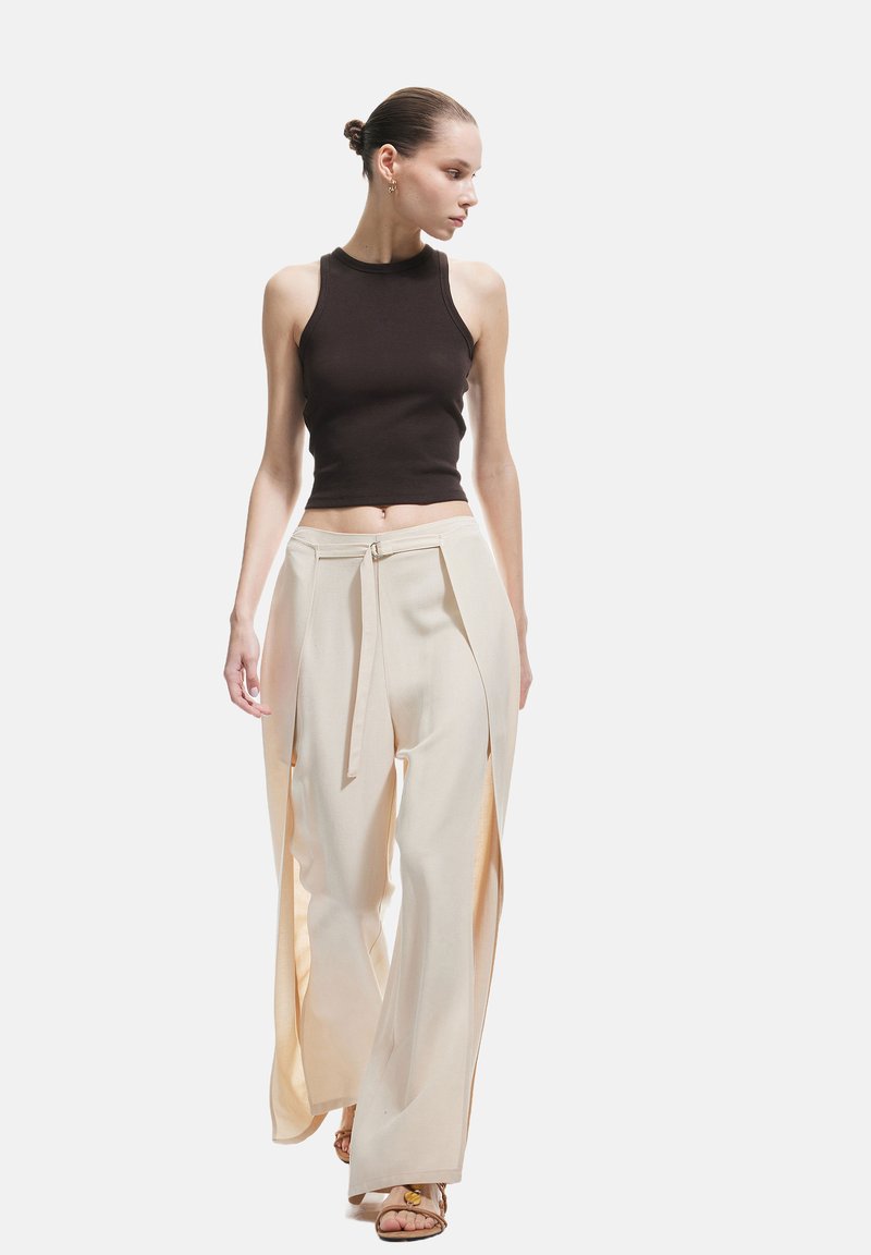 Schwarzes geripptes Halter-Crop-Top, kombiniert mit cremefarbenen weiten Hosen mit seitlichen Schlitzen und einem gebundenen Taillendetail, präsentiert vor einem einfarbigen Hintergrund.