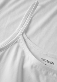 Vitt bomullslinne med tunna band, med rundad halsringning och etiketten "TWO SOON" tryckt i grått på innerfogen.