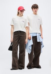 Δύο μοντέλα φοράνε λευκά t-shirts με λογότυπο και καφέ παντελόνια με φαρδύ πόδι; το ένα κρατά μια μαύρη τσάντα και φορά κόκκινο καπέλο, το άλλο δένει ένα μπλε ριγέ πουκάμισο γύρω από τη μέση.