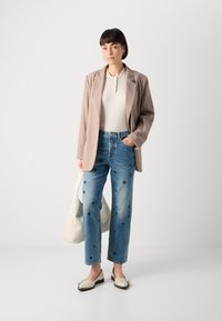 Lys beige blazer over en hvit ribbestrikket polo, blå broderte jeans og kremfargede leiligheter. Holder en hvit veske. Enkle designelementer er synlige.
