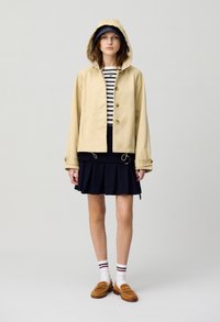 Veste de pluie khaki avec capuche, fermeture à boutons et manches longues. Portée sur un haut rayé et une jupe plissée bleu marine, assortie avec des mocassins beiges.