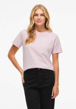 Szőke nő világos rózsaszín rövid ujjú pólóban és fekete nadrágban, egyik kezét a csípőjén nyugtatva, semleges háttér előtt áll.