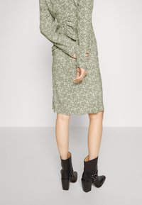 Grünes Blumenmusterkleid mit langen Ärmeln, figurbetontem Design und ausgestelltem Saum, kombiniert mit schwarzen klobigen Stiefeletten mit Absatz.