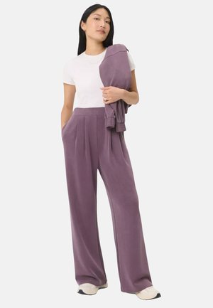 Vrouw draagt een wit T-shirt en hoge taille wijde mauve broek, houdt bijpassend mauve jasje over haar schouder, staand op een witte achtergrond.