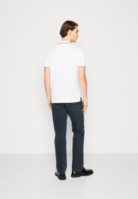 Witte polo met korte mouwen en een zwarte rand, gecombineerd met donkerblauwe jeans en zwarte schoenen; te zien vanuit de achterkant.