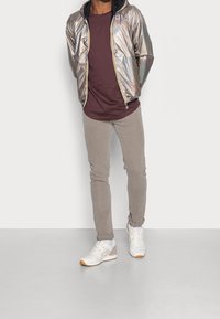 Man in een glanzende zilveren zip-up jas over een bordeauxrood shirt, lichtgrijze broek en witte sneakers, staande tegen een eenvoudige witte achtergrond.