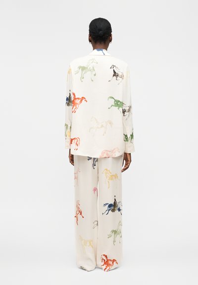 Henkilö pukeutunut kermaan vivahtavaan löysään asuun, jossa on värikkäitä hevoskuvioita, seisoo selin yksiväristä vaaleaa taustaa vasten.