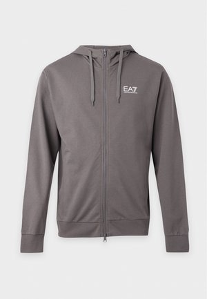 Homme debout portant un sweat à capuche gris zippé avec logo, pantalon noir et baskets noires sur fond blanc uni.