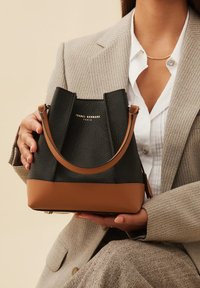 Black leather bucket bag with brown base, textured finish, short handle, and gold logo. Structured design with a spacious interior.