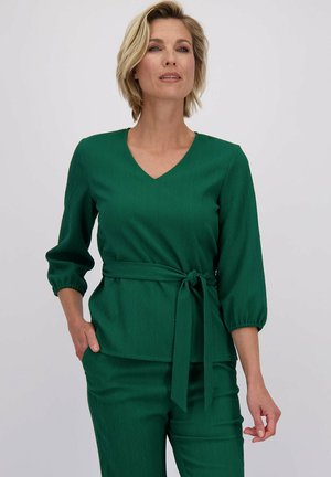 Haut vert en V avec des manches trois-quarts, taille cintrée par un lien, et tissu texturé présentant un motif en zigzag subtil.