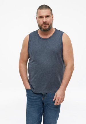 Mężczyzna z brodą i związanymi włosami, ubrany w niebieską koszulkę bez rękawów i jeansy, stoi z jedną ręką w kieszeni na białym tle.