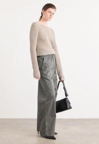 Beige gebreide trui met lange mouwen, gecombineerd met olijfgroene wijde broek. Model houdt een zwarte handtas vast en draagt puntige zwarte hakken.