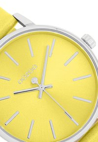 Montre analogique jaune avec accents argentés, dotée d'un cadran lisse, de marqueurs en bâton et d'un bracelet en cuir mince. Nom de la marque "oozoo" affiché.