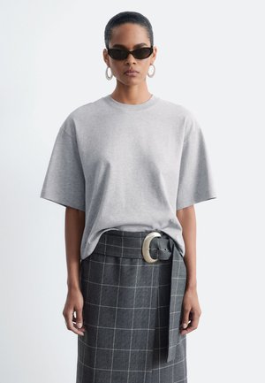 Femme portant des lunettes de soleil noires, des boucles d'oreilles créoles argentées, une chemise oversize à manches courtes gris clair et une jupe grise à carreaux avec une large ceinture et une grande boucle.