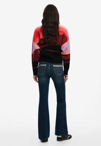 Maglione multicolore con motivo astratto, fondo nero, abbinato a jeans svasati blu scuro con dettagli delle tasche azzurri.