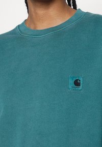 Sudadera de algodón color azul verdoso con cuello redondo y pequeña etiqueta con el logo de Carhartt en negro y azul en el pecho izquierdo. Textura suave con puños acanalados.