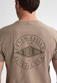 Ruskea lyhythihainen t-paita, jossa on tekstuuri. Edessä tummanvihreä ympyräkuvio, jossa lukee "DENIM DRIVEN PETROLIND FLORIDA U.S.A. '89."
