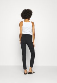 Jean noir coupe slim avec une taille mi-haute, doté de poches arrière et d'une texture lisse, porté avec un haut blanc sans manches et des sandales.