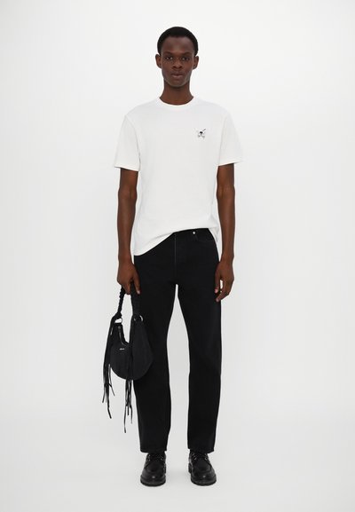 White T-shirt with a small black butterfly print on the chest, paired with black high-waisted jeans and a black shoulder bag with fringe detail.