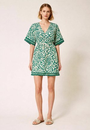 Donna che indossa un vestito corto verde e bianco con motivo, bordatura in pizzo e coulisse in vita, in piedi con espressione neutra e sandali beige.