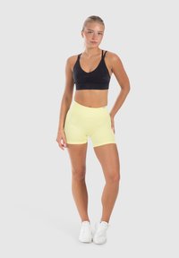 Zwarte sportbeha met een V-hals en een geplooid detail aan de voorkant, gecombineerd met hoge gele shorts. Model staat in witte sportschoenen.