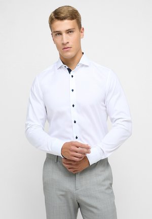 Camicia bianca a maniche lunghe con bottoni blu navy e dettagli del colletto, caratterizzata da una consistenza liscia e un design aderente. Abbinata a pantaloni grigi chiari.