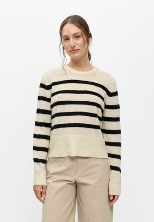 Femme aux cheveux châtain clair portant un pull beige avec des rayures horizontales noires et un pantalon beige clair, debout devant un fond blanc.