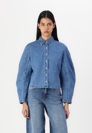 Cropped denim jacket in medium blue, featuring a button-down front, large sleeves, and visible stitching details. Styled with matching denim pants.