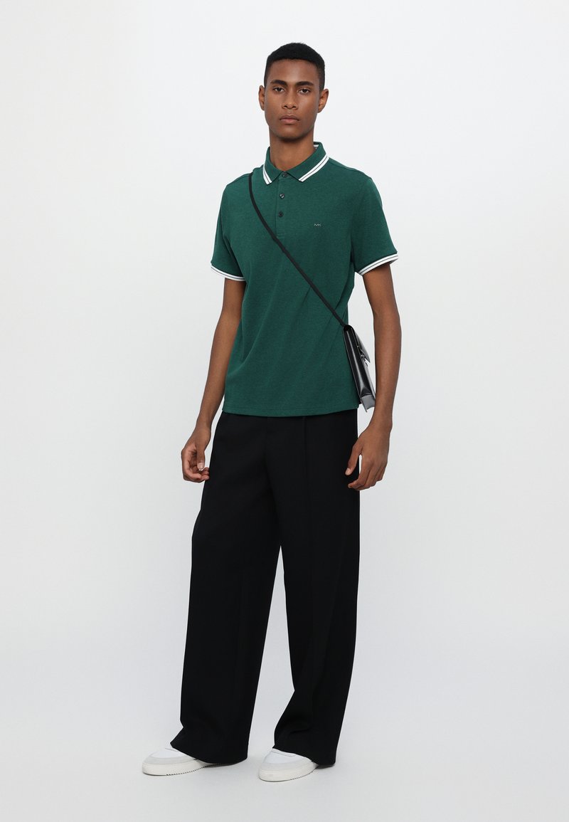 Dunkelgrünes Polohemd mit weißen Kragen- und Ärmelstreifen, kombiniert mit lockeren schwarzen Hosen und weißen Sneakers, während eine kleine schwarze Tasche gehalten wird.