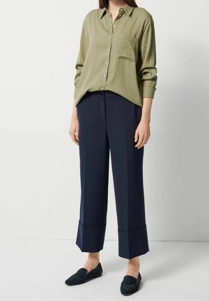 Kvinde iført olivengrøn skjorte med knapper, marineblå vidde-bukser med opsmøg og sorte slip-on loafers, stående mod en hvid baggrund.