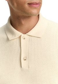 Nahaufnahme einer Person, die ein creamfarbenes, strukturiertes Strick-Polo-Shirt mit einer Knopfleiste mit zwei Knöpfen und einem Kragen trägt.