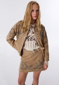 Tan suède jasje en rokset met metalen versieringen, gelaagd over een gebreide top, met franjes en een ceintuur in de taille.